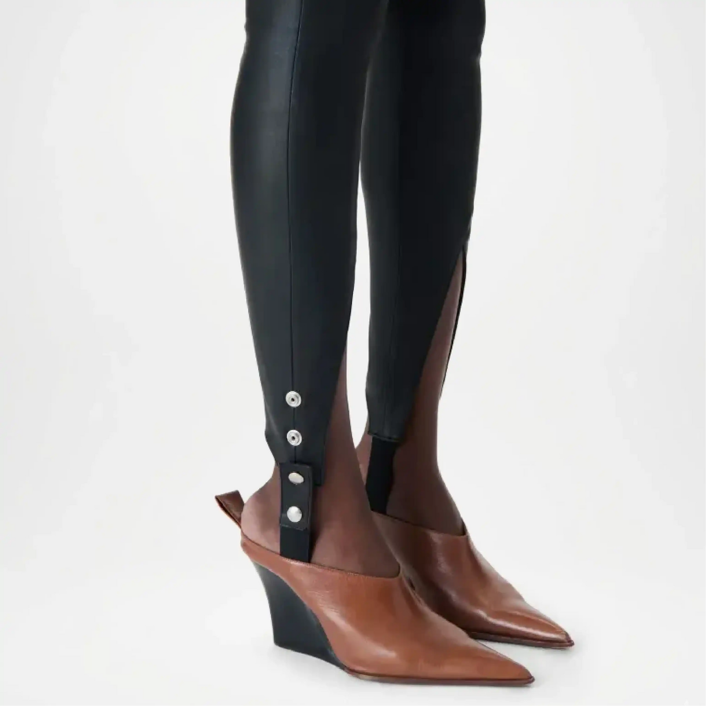 Brown pointed-toe shoes with black wedge soles worn with black leggings on a light gray background
