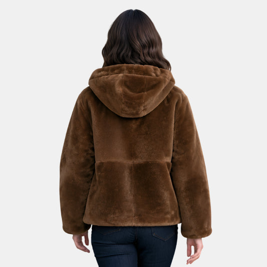 A woman viewed from the back wearing a brown plush faux-fur hooded jacket and dark blue jeans against a light gray studio background.