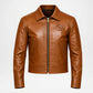 A brown leather aviator-style jacket with a front zipper, pointed collar, and a circular embroidered patch on the left chest, shown against a plain light gray background.