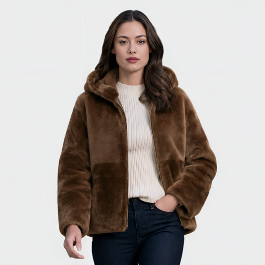 A woman modeling a brown faux fur jacket with a hood, worn over a cream ribbed sweater and dark blue jeans, against a light gray studio background.